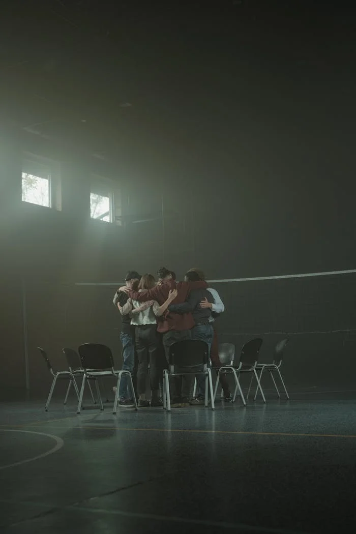 A diverse support group hugs in a dimly lit room, fostering unity and emotional support.